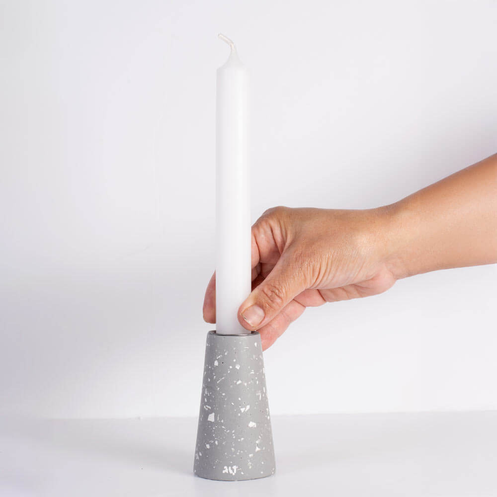A hand holding a white taper candle in a grey taper candle holder with white terrazzo, set against a plain white background.