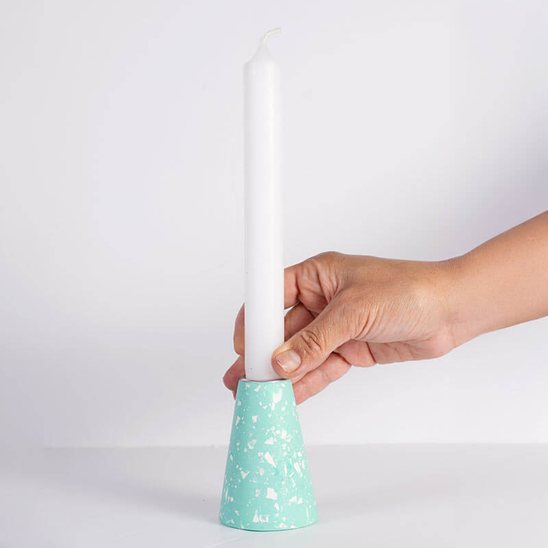 Hand holding a white taper candle and a mint green and white terrazzo-patterned taper candle holder with a silver-toned top, shown on a white background