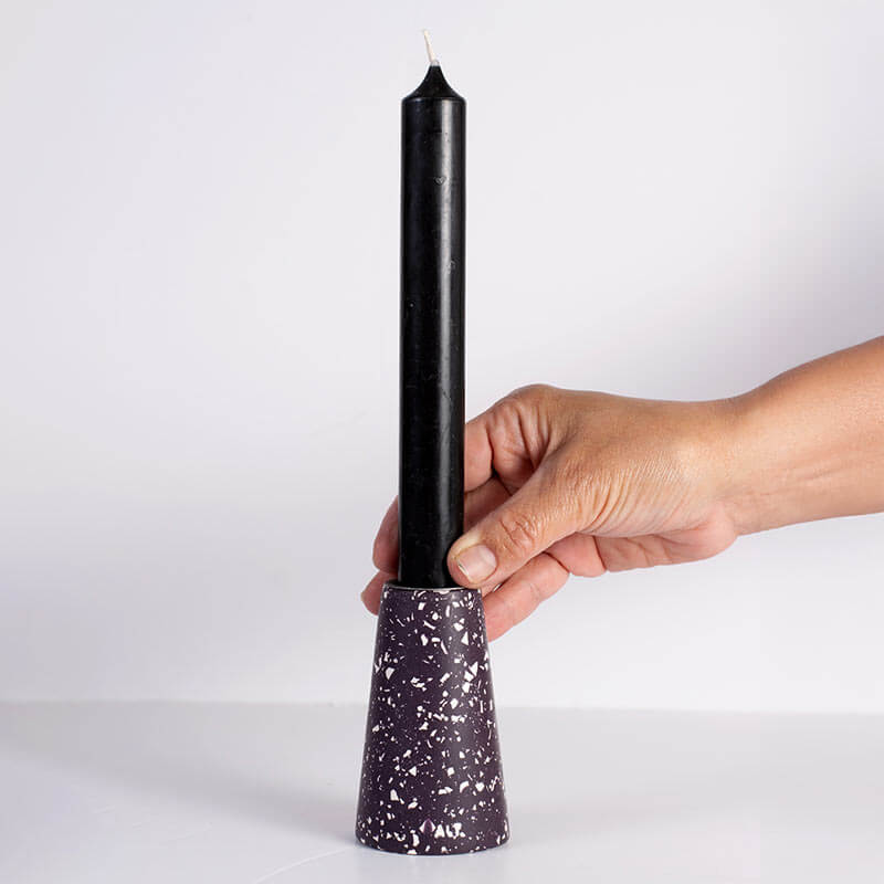 A hand holding a black taper candle in a white speckled dark purple taper candle holder against a plain white background.