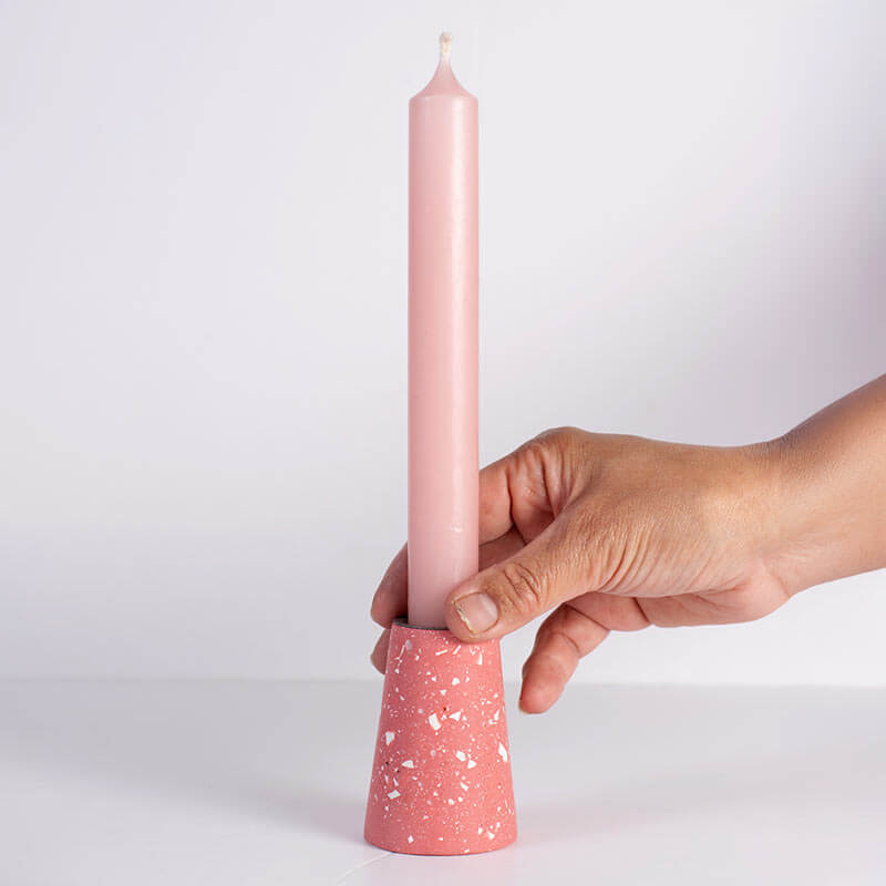 A hand holding a tall light pink taper candle in a salmon pink-coloured cone-shaped taper holder with white speckles, set against a plain light gray background.