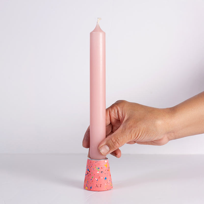 A hand holding a tall, slim pink taper candle in a pink rainbow terrazzo taper candle holder against a clean white background.