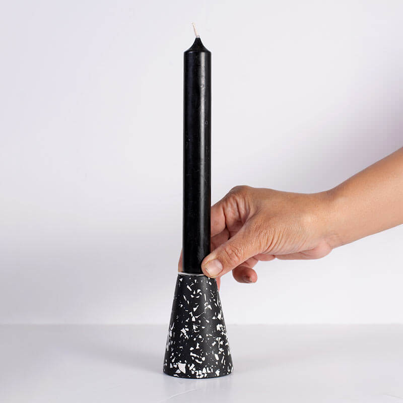A hand holding a black taper candle in a black and white speckled taper candle holder against a plain white background.