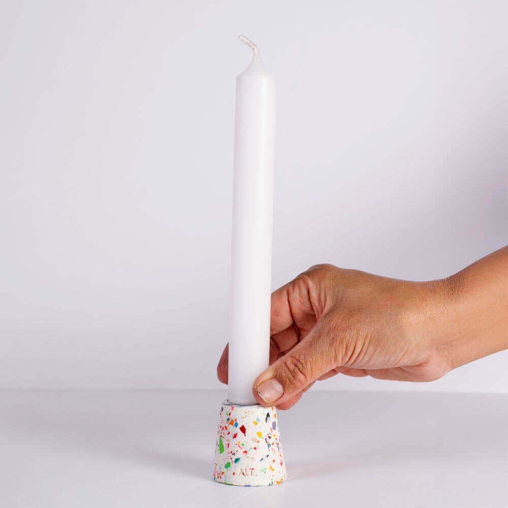 A hand holding a white taper candle in a white and rainbow terrazzo patterned taper candle holder with a silver-toned top, shown on a white background