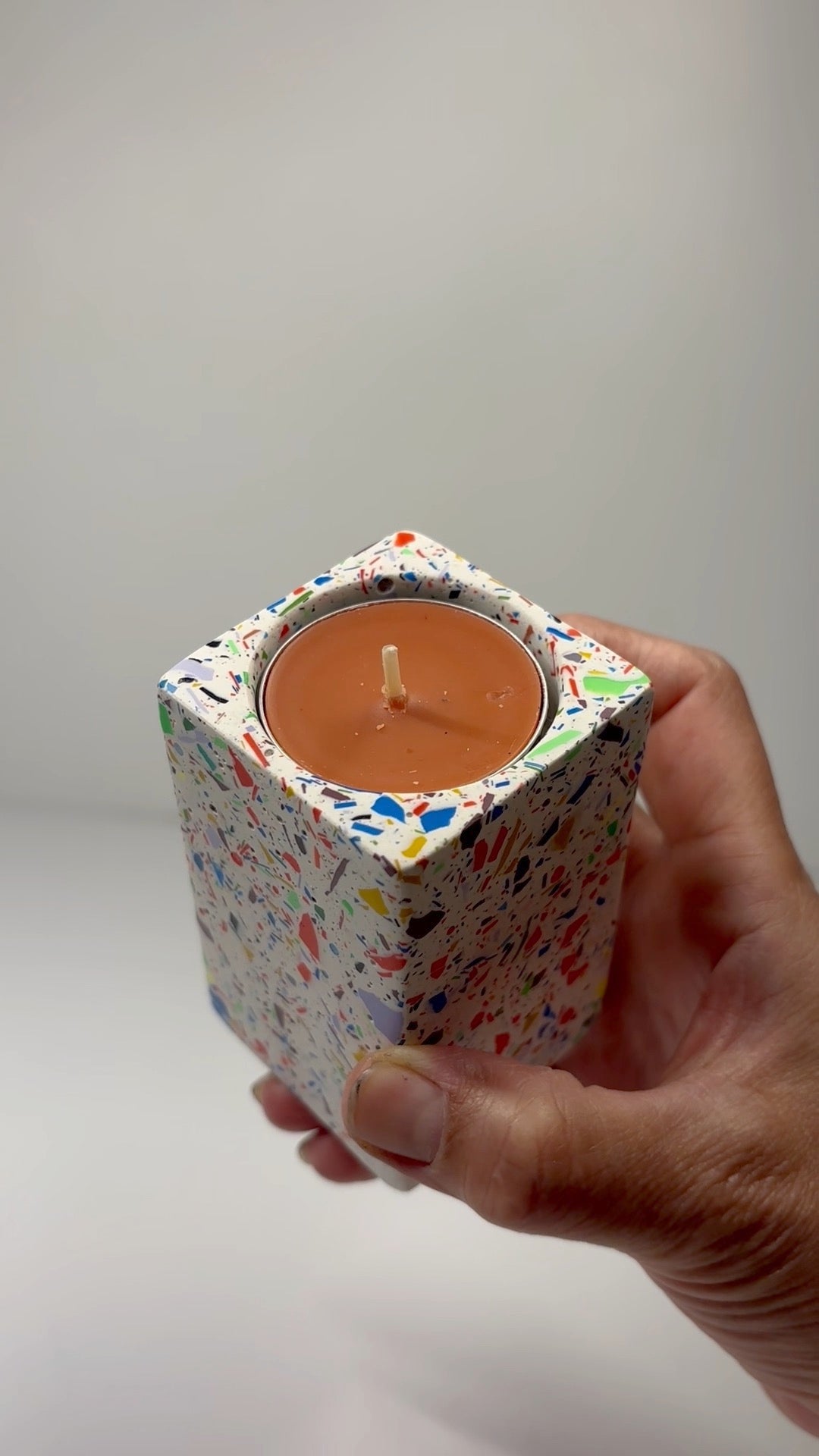 Hand holding a colourful rainbow rectangular shaped tall tea light candle holder with a tea light candle inside on a plain background
