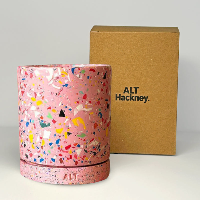 Pink terrazzo-patterned planter with a drainage hole and a water tray on a book  next to a brown box labeled 'ALT Hackney' on a light gray background.