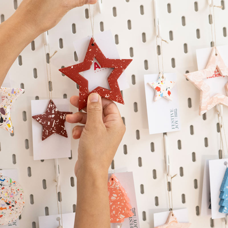 A hand holding a red star-shaped ornament with 'ALT Hackney' festive card on a pegboard with other decorative items.