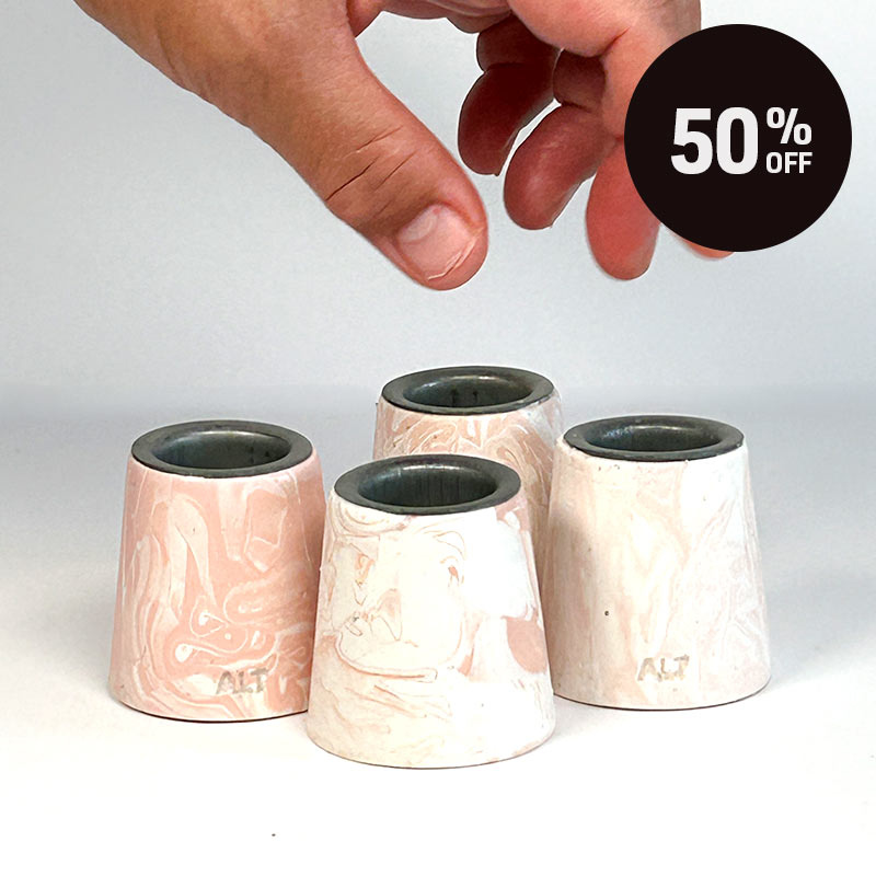 Four small tapered candle holders with a salmon pink and white marbled pattern, arranged on a white surface. A hand is hovering above them, and a black circular '50% OFF' sale badge is in the top-right corner.