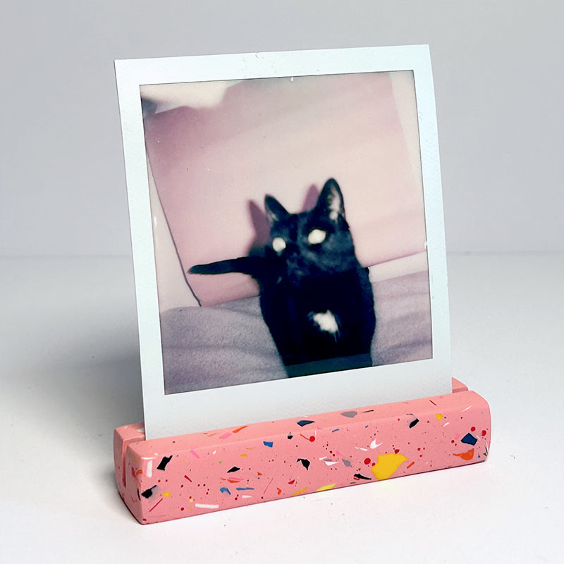 A pink terrazzo-patterned photo stand holding a Polaroid-style instant photo of a black cat, photographed on a clean white background.