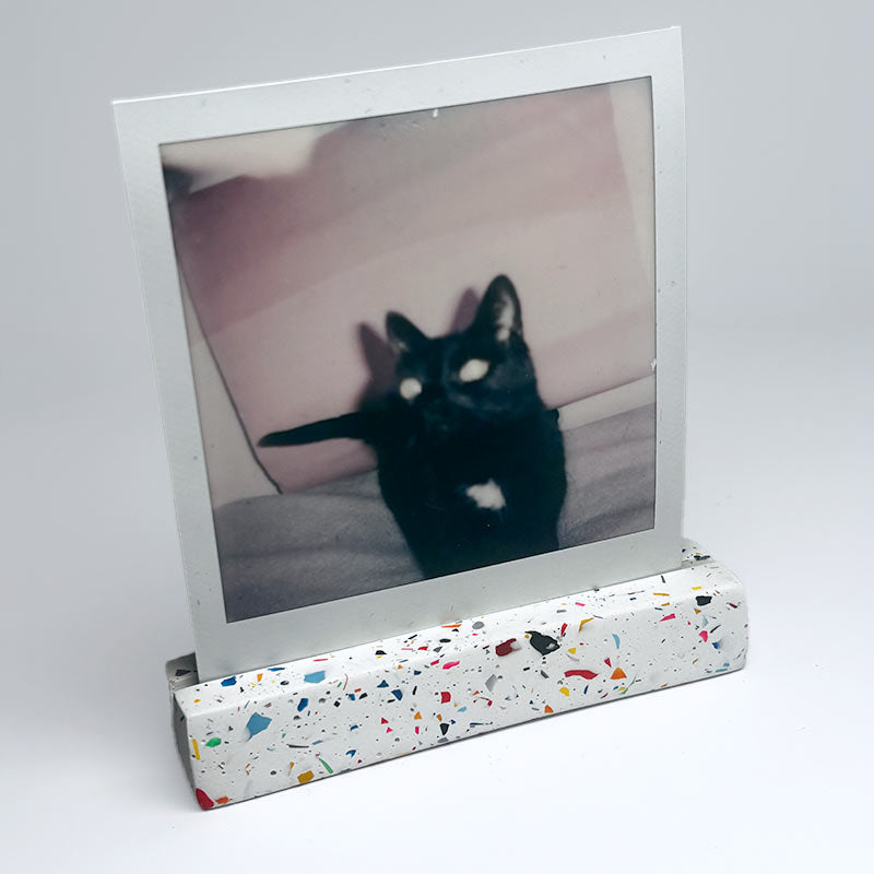 A raibbow terrazzo-patterned photo stand holding a vertical instant photograph of a black cat, set against a plain light gray background.