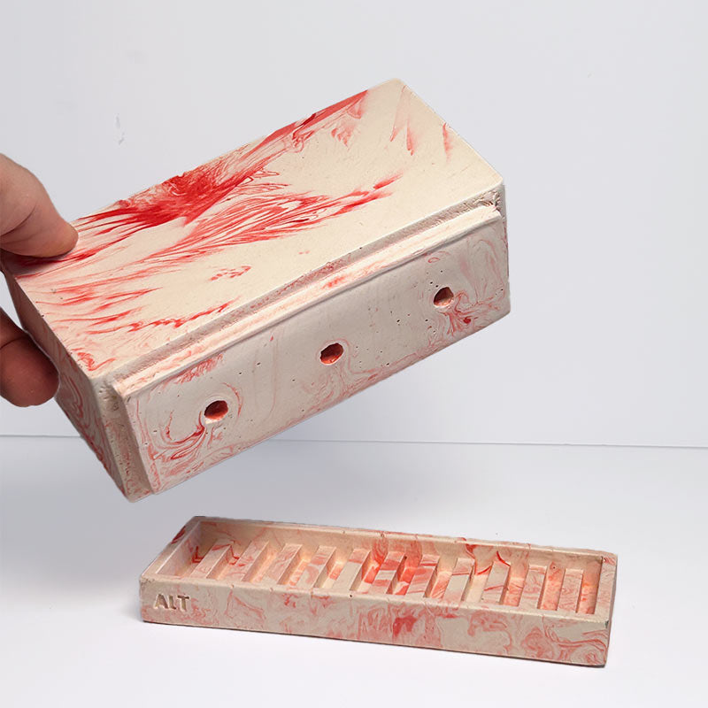 A hand holds a rectangular planter above a matching shallow tray with carved slots, both showing a marbled off-white surface with red painted streaks. The background is a clean white studio backdrop.