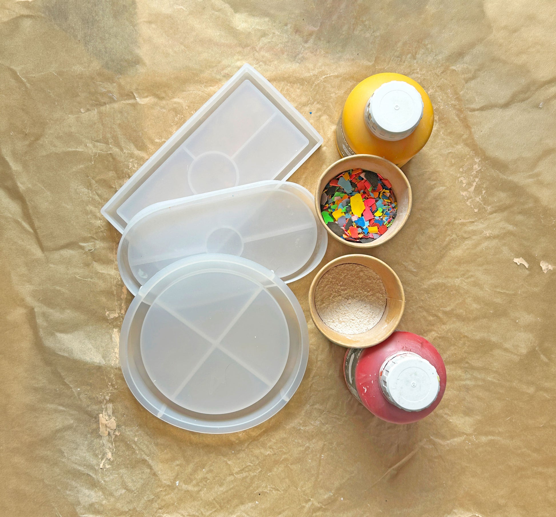 3 differently shaped clear silicone moulds on brown paper, with small containers of colourful terrazzo eco-resin and two bottles of pigment.