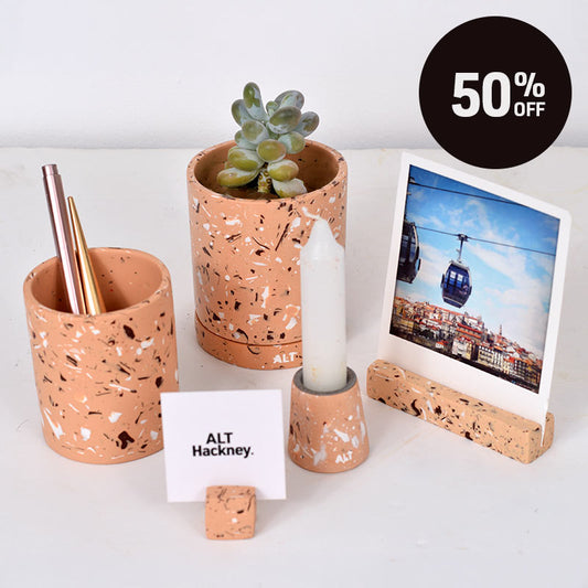 A beige, brown and white terrazzo plant plant pot with a small succulent, next to a matching pen pot,  photo holder with a polaroid photo of a cable car city scene and another small photo holder with ALT Hackney business card  and a small taper candle holder with a small white taper candle in. A black circular badge in the top right reads '50% OFF'.