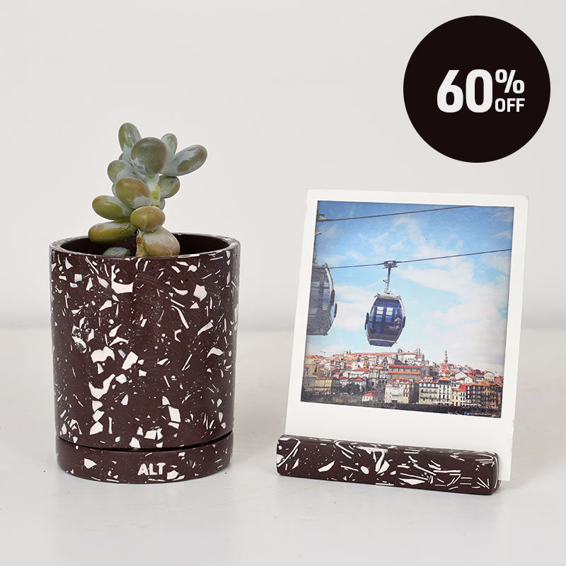 A brown and white terrazzo plant plant pot with a small succulent, next to a matching photo holder with a polaroid photo of a cable car city scene. A black circular badge in the top right reads '60% OFF'.