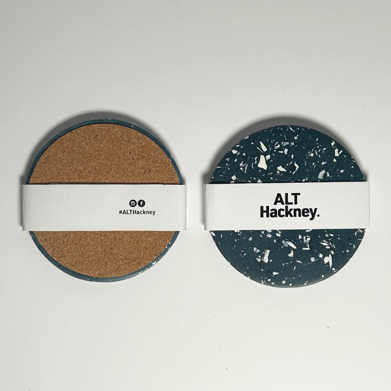 Two round coasters with white paper bands across the middle, displayed side by side on a light gray background. The left pad has a natural cork texture, and the right pad has a dark navy and white terrazzo pattern.