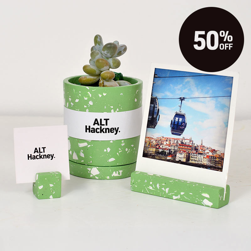 A green and white terrazzo plant plant pot with a small succulent, next to a matching photo holder with a polaroid photo of a cable car city scene and another small photo holder with ALT Hackney business card . A black circular badge in the top right reads '50% OFF'.
