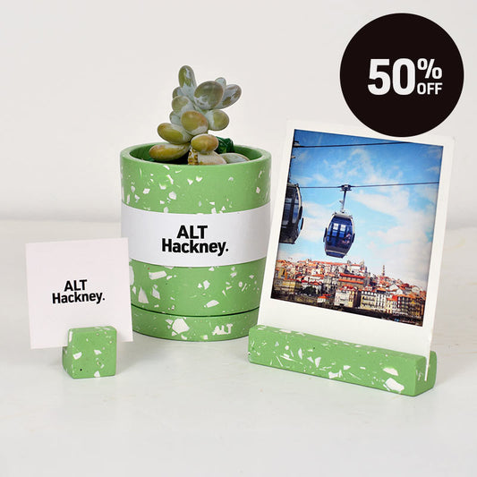 A green and white terrazzo plant plant pot with a small succulent, next to a matching photo holder with a polaroid photo of a cable car city scene and another small photo holder with ALT Hackney business card . A black circular badge in the top right reads '50% OFF'.