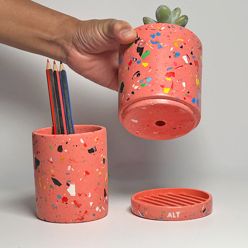A Pink rainbow coloured pencil holder with a colourful speckled pattern, shown with a hand holding the planter, a pencil cup containing coloured pencils, and the water tray placed on a white surface.