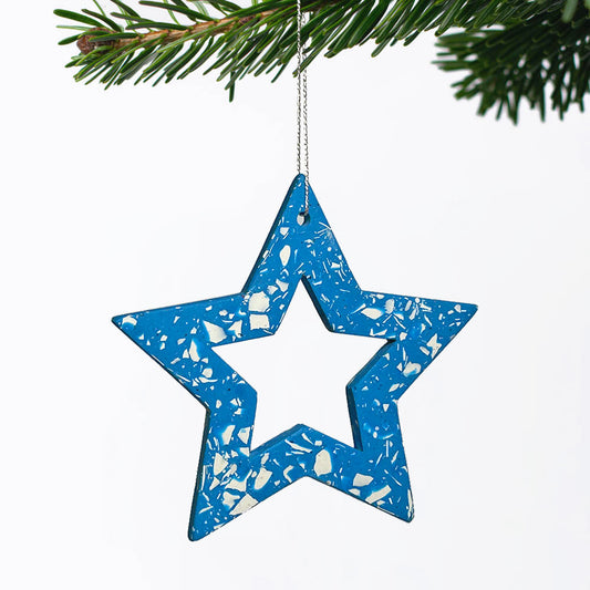 A blue star-shaped Christmas ornament with a white terrazzo speckled pattern, hanging from a silver string on a pine branch against a white background.