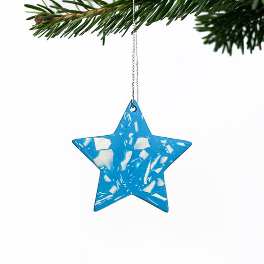 A star Christmas ornament in bright blue with a white marbled pattern, hanging from a silver string under pine tree branches against a white background.