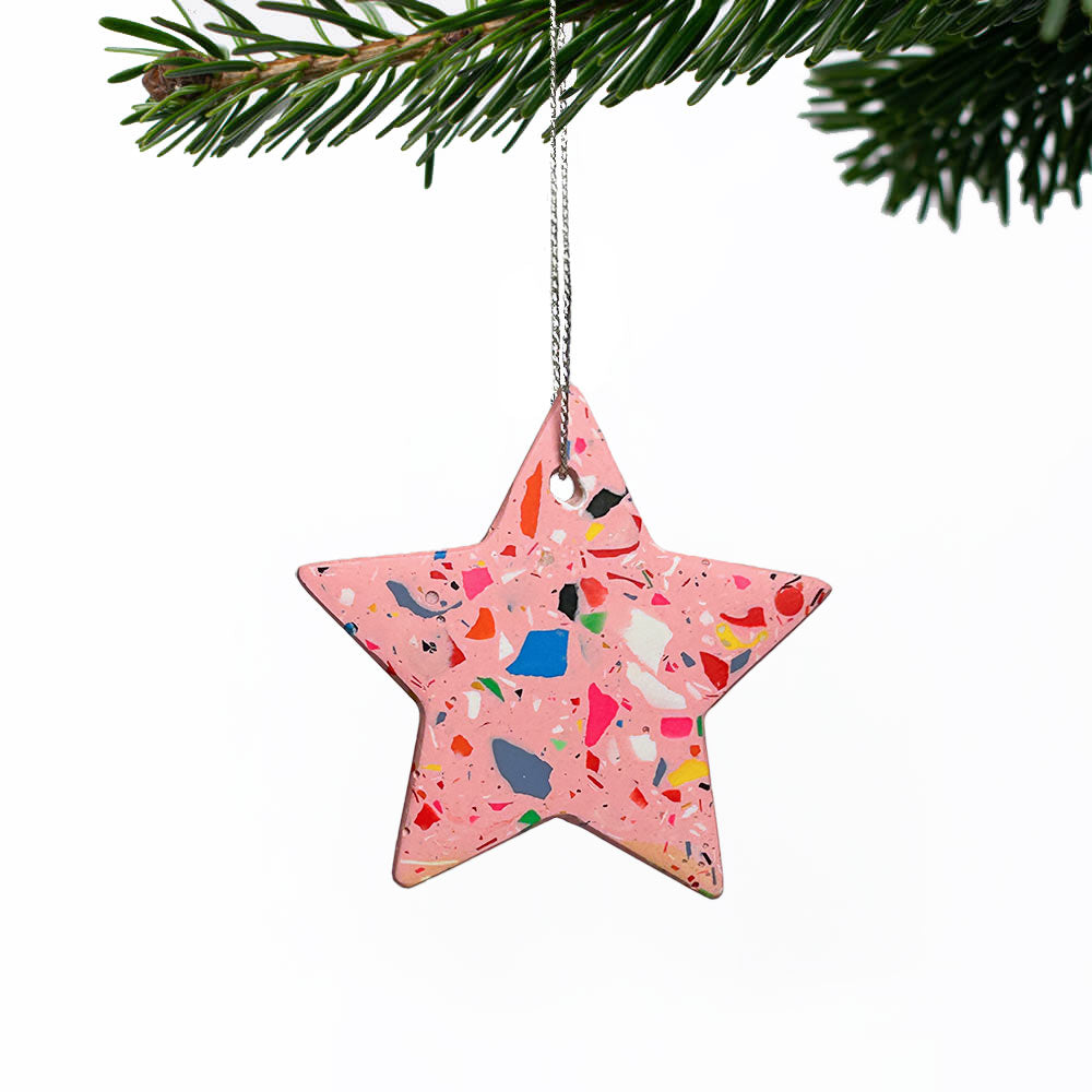 A pink star-shaped Christmas ornament with a terrazzo pattern of multicolored chips, hanging from a silver string from a pine branch against a white background.