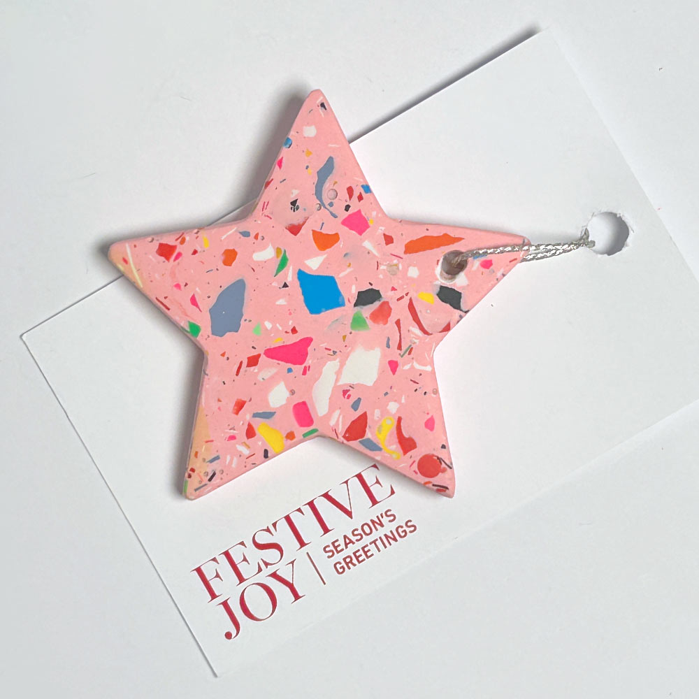 A pink terrazzo-patterned star-shaped ornament with a silver hanging loop, displayed on a white greeting card that reads 'FESTIVE JOY | SEASON'S GREETINGS'.