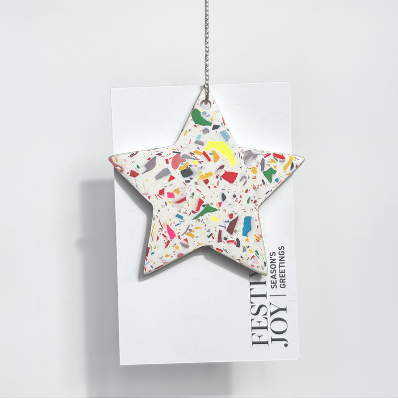 A star ornament with a coloうrful terrazzo pattern on a white background, hanging from a silver chain on a white greeting card that reads 'FESTIVE JOY SEASON'S GREETINGS'.