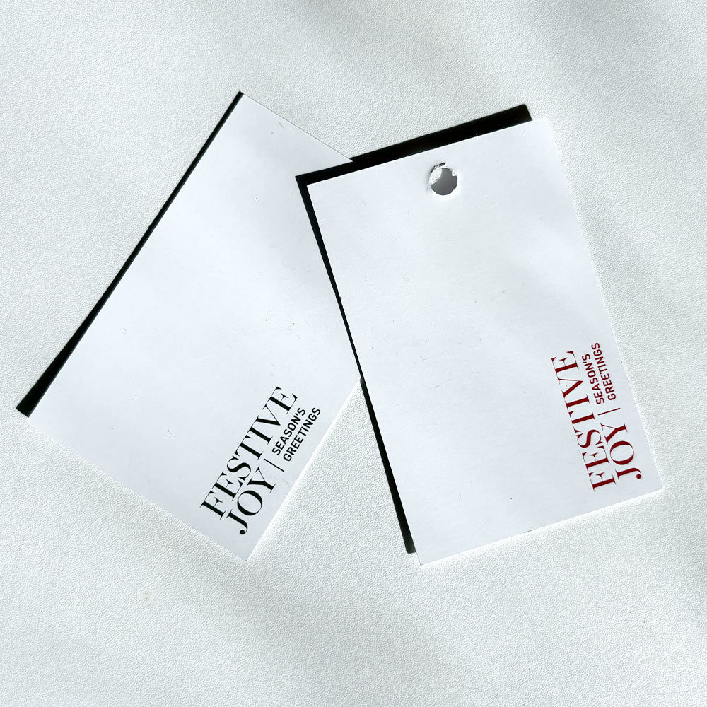 Two white paper gift tags with printed text reading 'FESTIVE JOY' and 'SEASON'S GREETINGS' on a light grey background.