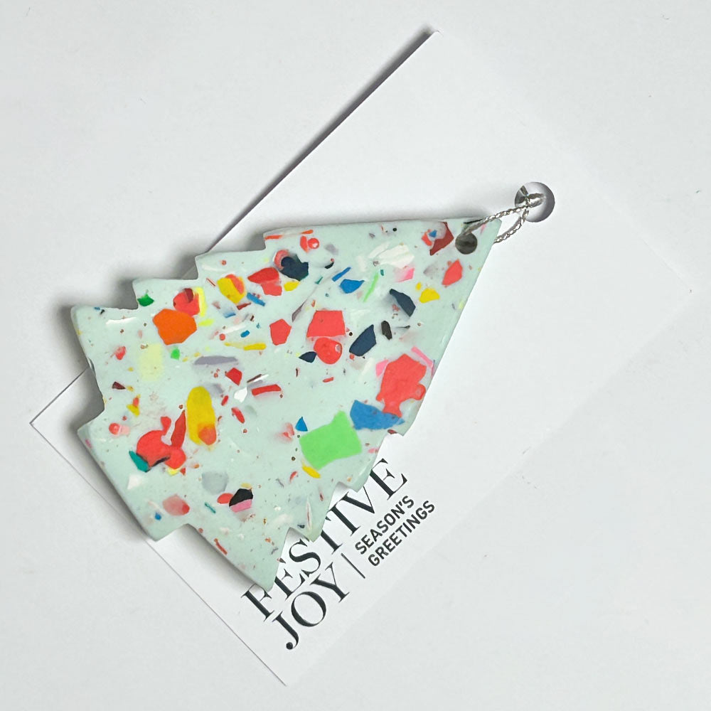 A tree ornament with a multicolor terrazzo pattern on a pale mint green base, displayed on top of a white greeting card that reads 'FESTIVE JOY | SEASON'S GREETINGS'.