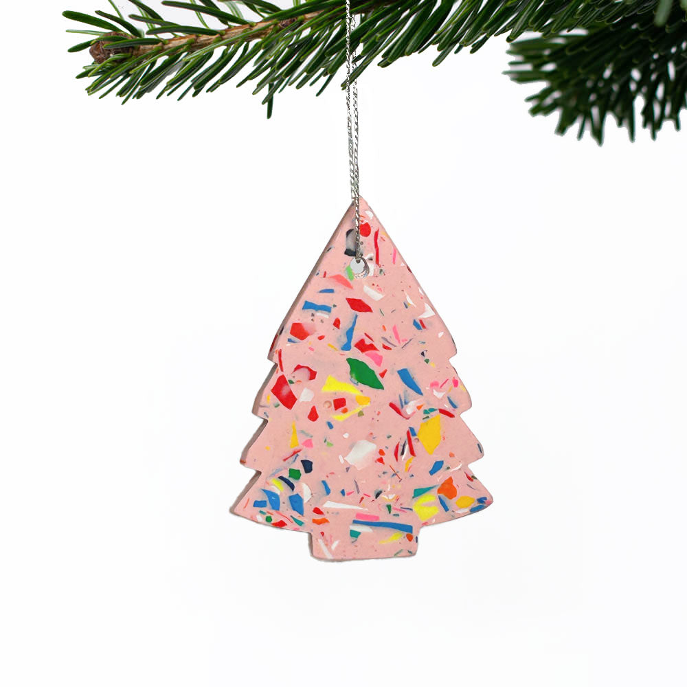 A pink Christmas tree ornament filled with multicoloured confetti, hanging from a silver string on a pine branch against a white background.
