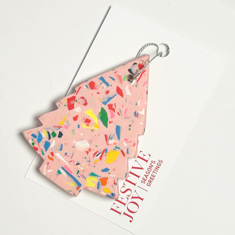 A pink Christmas tree ornament filled with multicoloured confetti, resting on a white greeting card that reads 'FESTIVE JOY SEASON'S GREETINGS'.