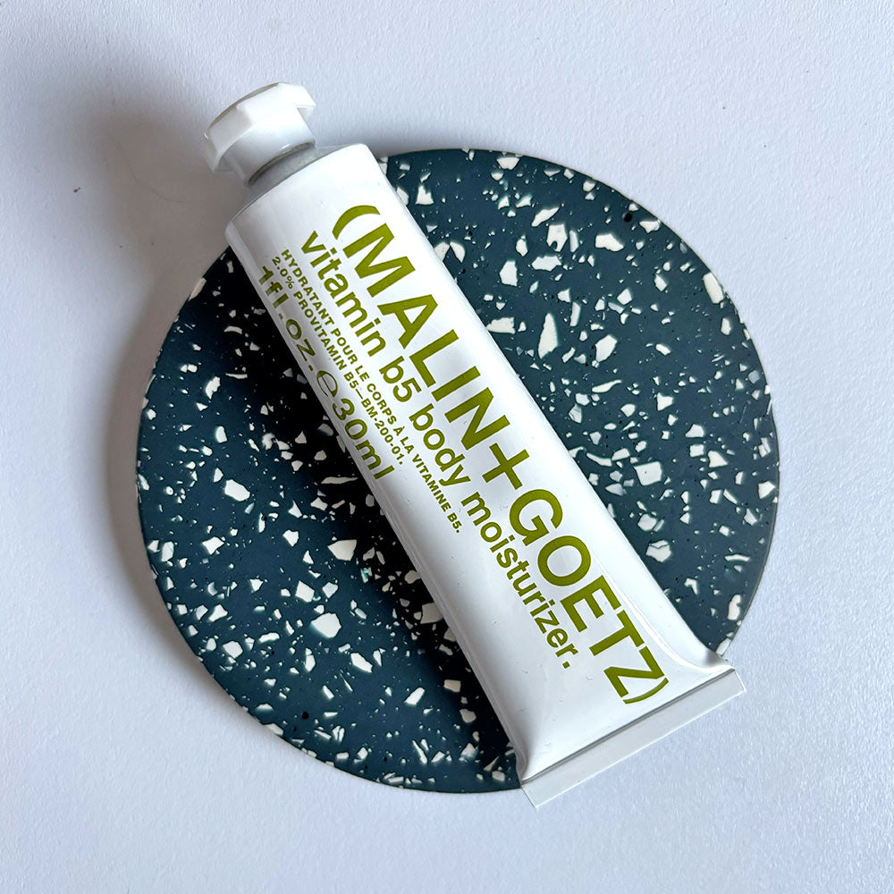 MALIN+GOETZ vitamin B5 body moisturizer tube on a navy terrazzo round coaster on a light gray background