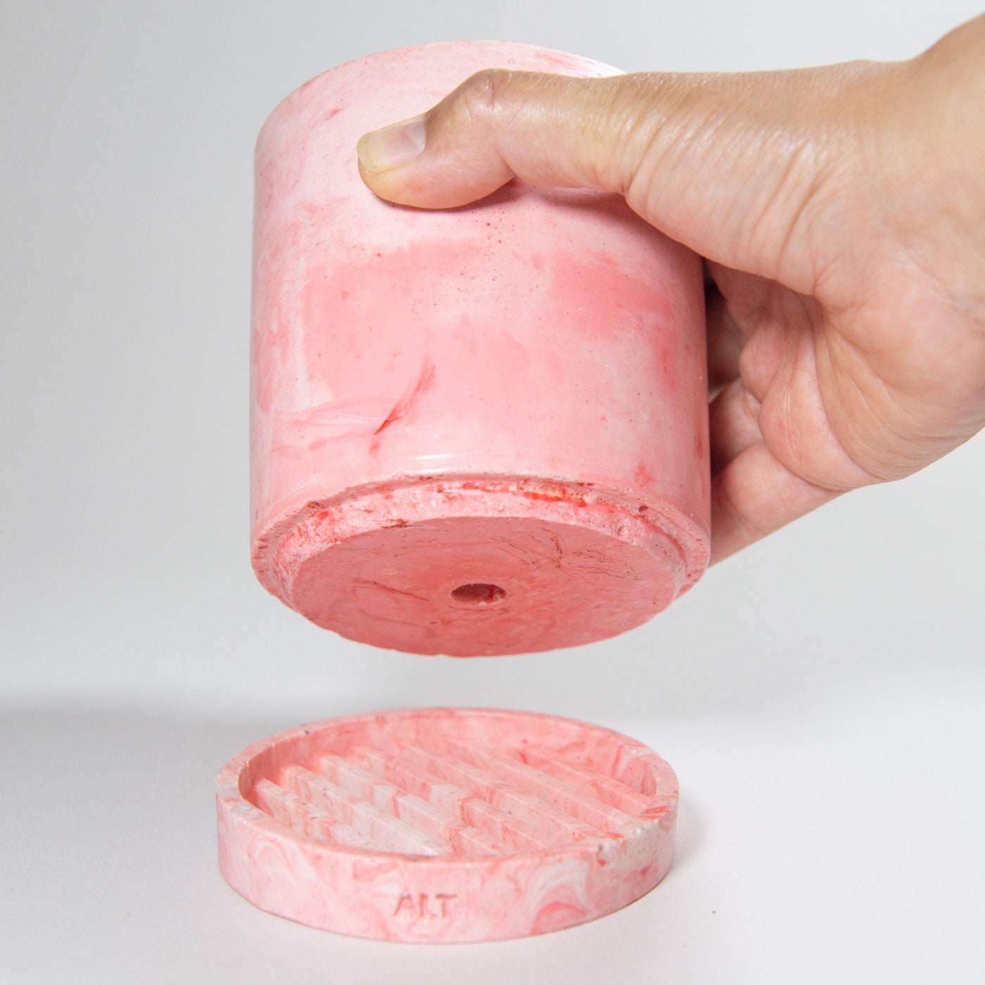 Pink marbled cylindrical plant pot with a drainage and a water tray with a hand holding it on a light gray background