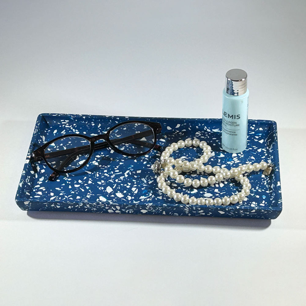 Navy blue speckled tray with glasses, pearls, and a bottle of serum on a white background