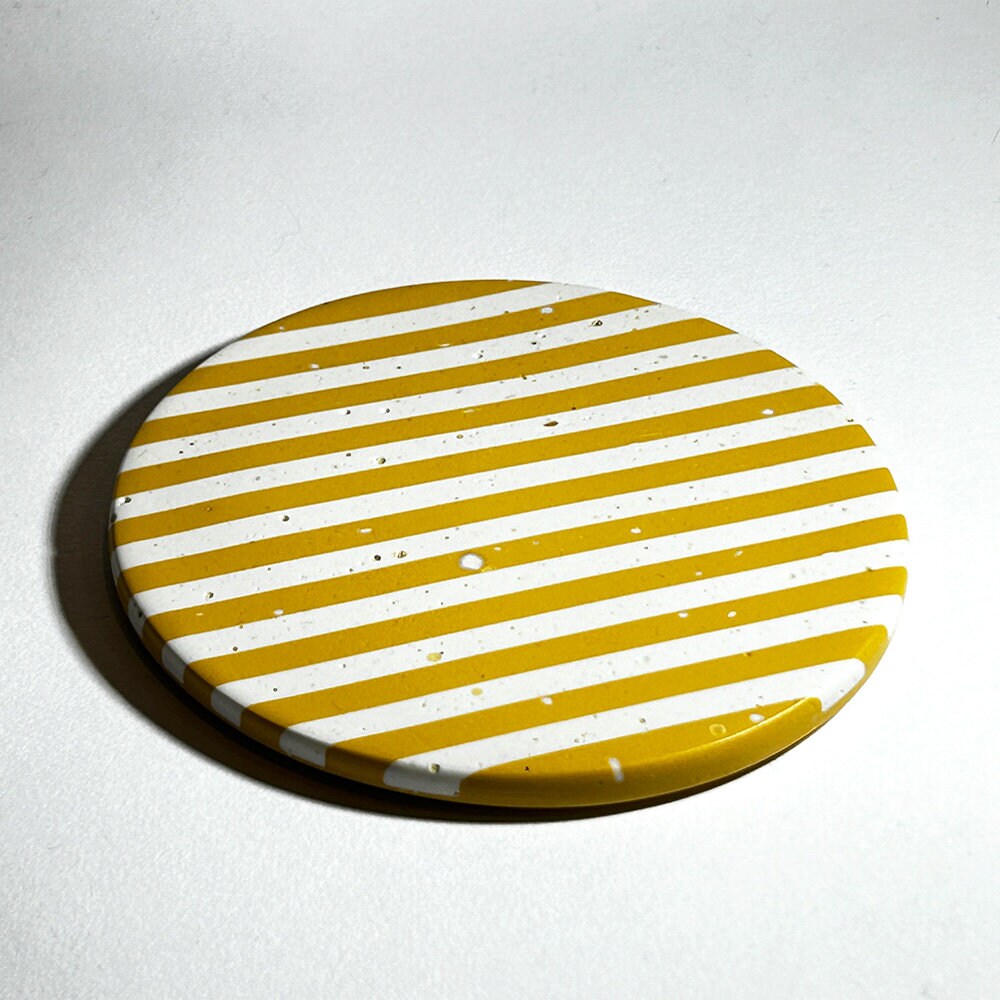 Round coaster with yellow and white striped pattern on a light grey background