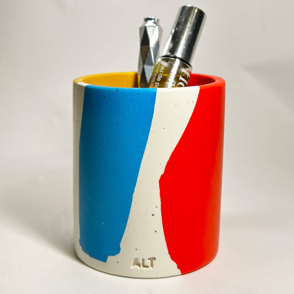 A pen pot with blue, red, and yellow colour block design with a pen and paper clips on a light background
