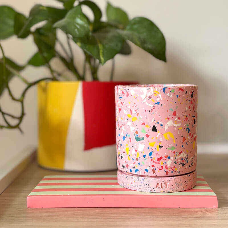 Pink terrazzo-patterned planter with a drainage hole and a water tray on a book on a striped surface with a plant in the background
