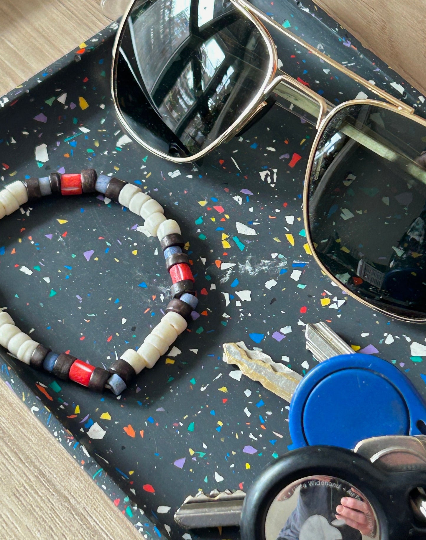 Sunglasses and a beaded necklace on a terrazzo tray with keys and a blue container.