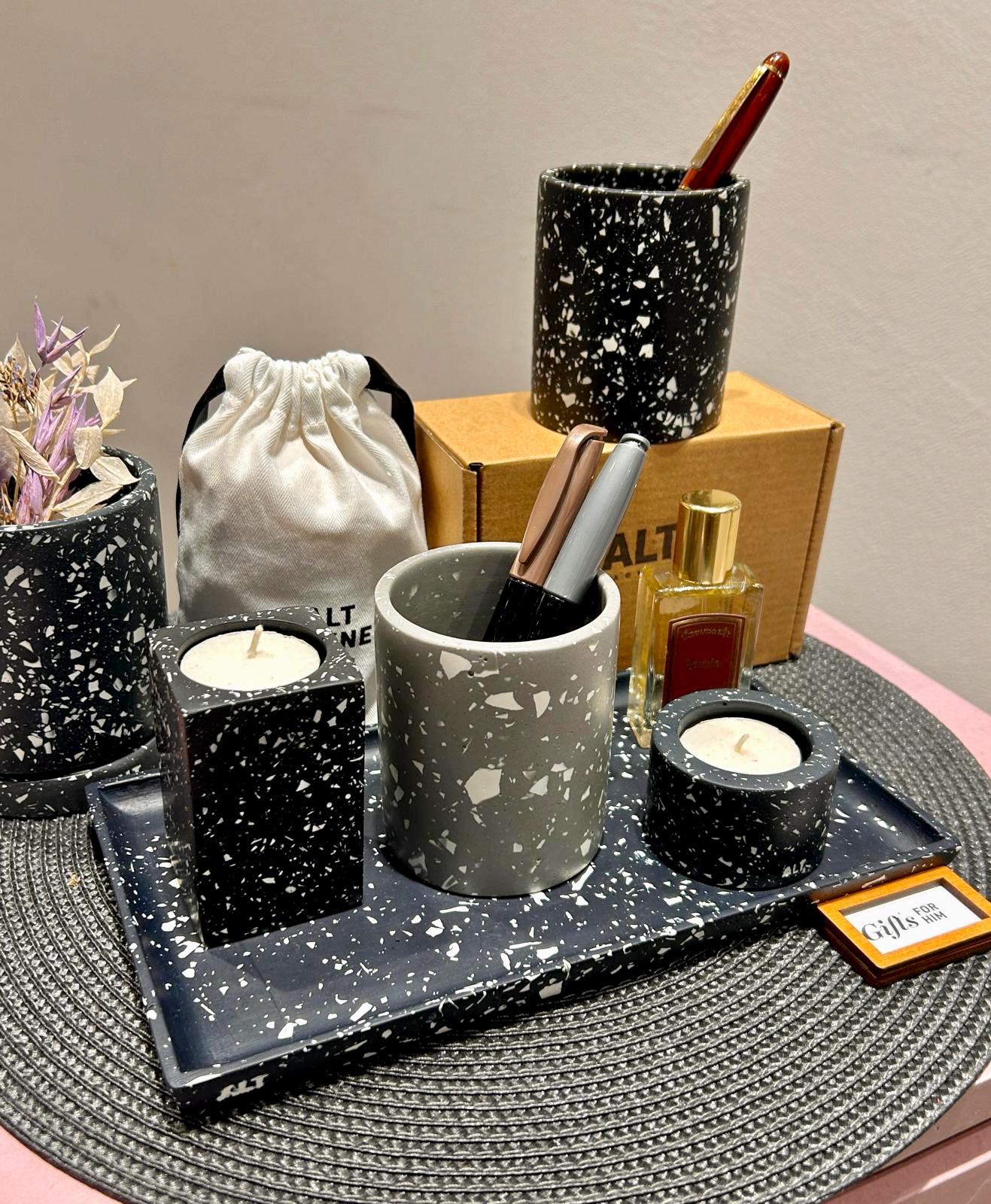 Various terrazzo homewares including pen pots, a tray, candle holders with candles and a plan pot on a textured surface.