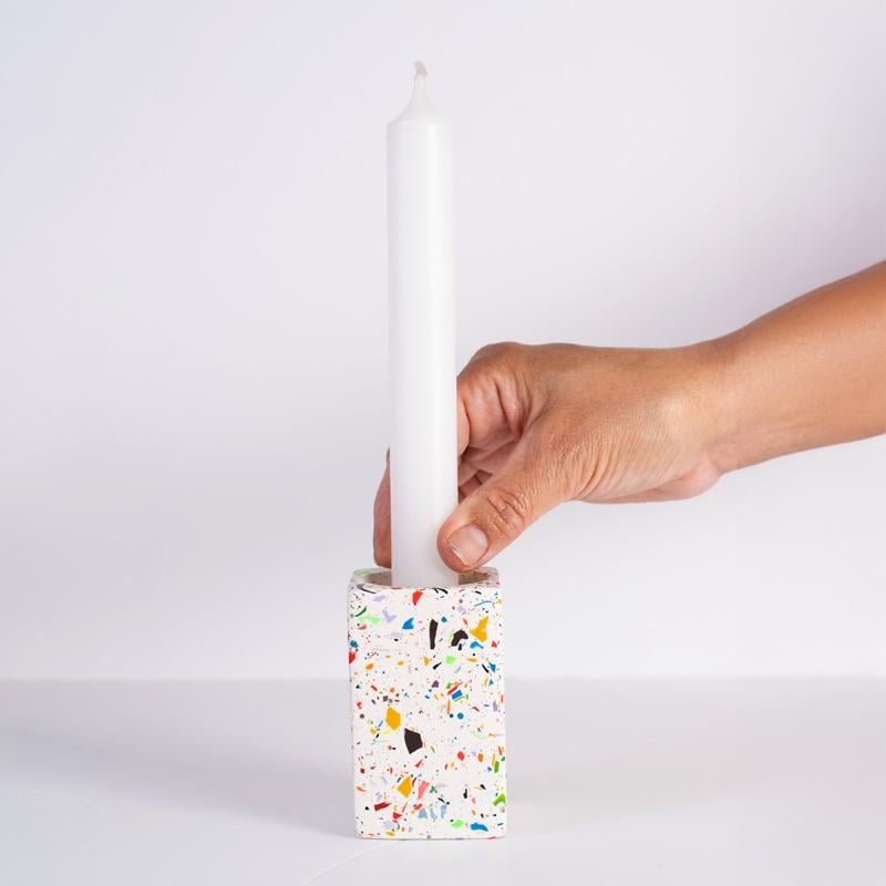 Hand holding a white candle in a colorful terrazzo-patterned holder against a light grey background