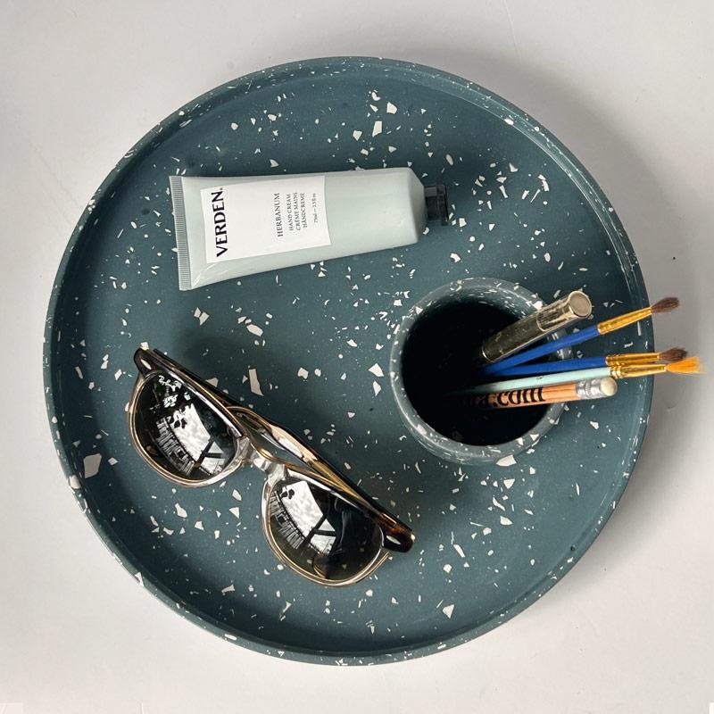 Round navy terrazzo tray with sunglasses, tube of cream, and makeup brushes on a light grey background