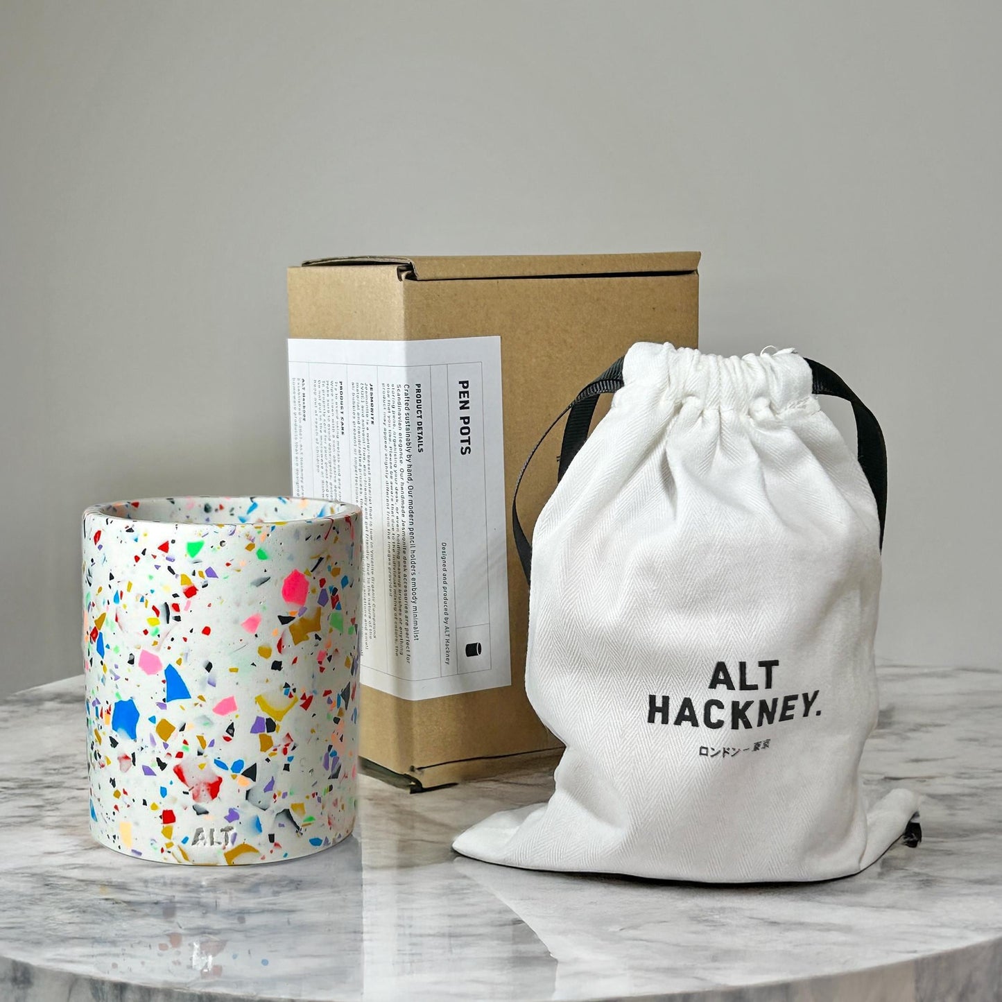 Colorful terrazzo-patterned candle, cardboard box, and white drawstring bag with 'ALT HACKNEY' branding on a gray surface.