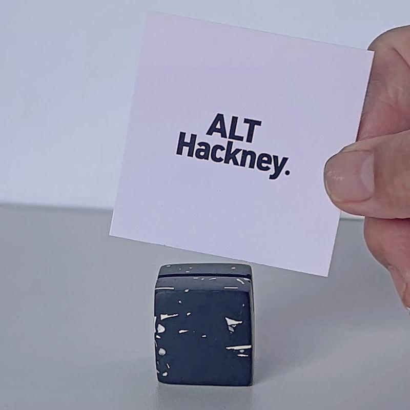 Hand holding a card with 'ALT Hackney.' text next to a blue cube on a light background
