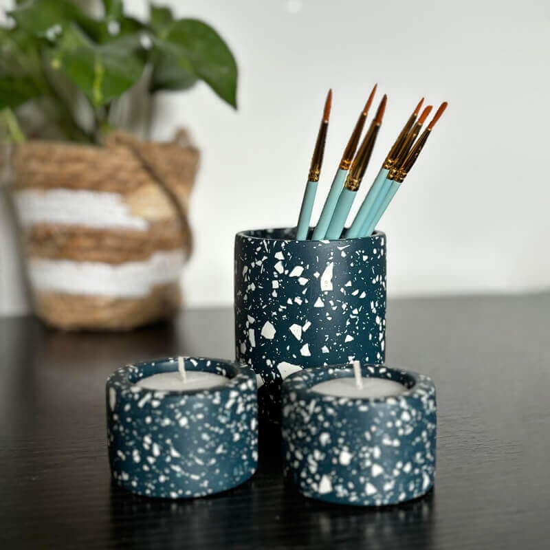 Various navy blue terrazzo candle holders with candles and a paintbrushes in a pen pot on a dark surface.