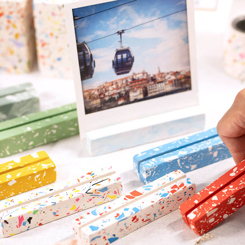 A close-up of a hand holding a long red-and-white terrazzo photo holder with several other photo holders in yellow, white, blue, and red arranged on a light surface. A printed photo postcard stands in the background showing a cable car scene.