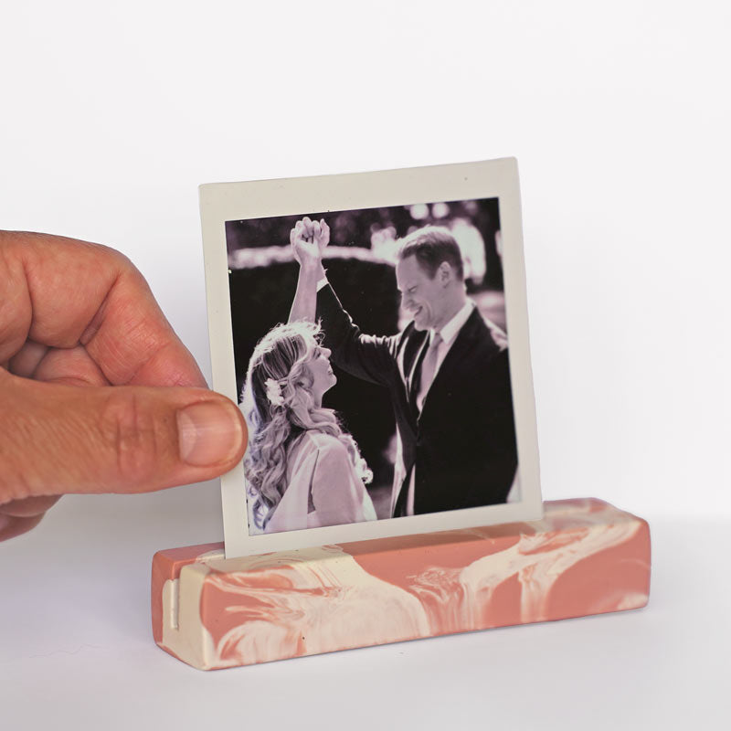 Hand holding a marble-patterned salmon pink and white photo holder with a wedding photo inside on a white background