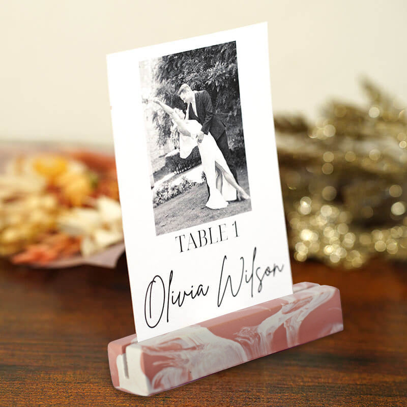 A personalised wedding table number card on a pink and white marble wedding place name card hodler, set on a wooden table with a blurred background.