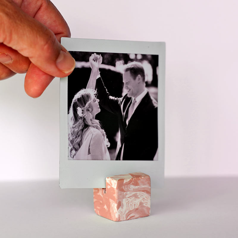 Hand holding a polaroid photo of a couple on a marble small square salmon pink photo holder against a white background