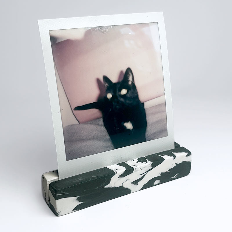 A dark green and white marble effect rectangular photo holder displaying a printed instant photograph of a black cat against a pink background, set against a clean light grey studio backdrop.
