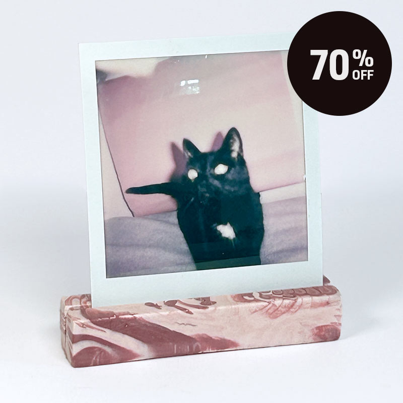 A square instant photo with a white border stands upright on a terracotta red with salmon pink marble effect rectangular display photo stand against a plain light grey background with a black '70% OFF' promotional badge in the top right corner.