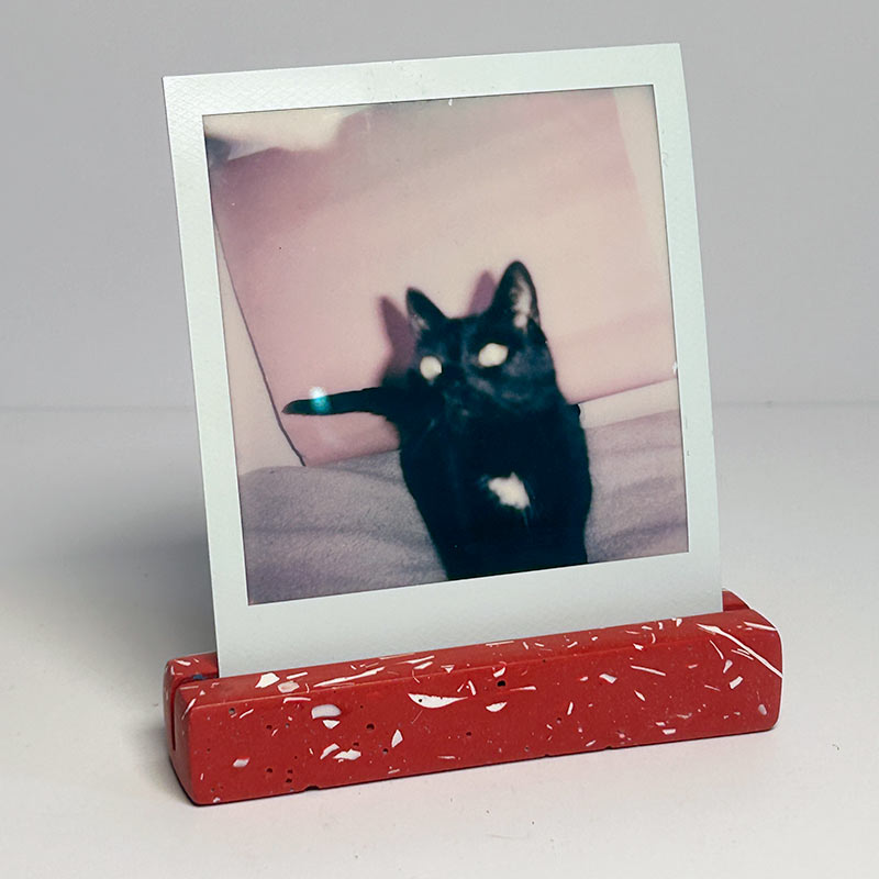 A square instant photo with a white border stands upright on a terracotta red speckled rectangular display photo stand against a plain light gray background. 